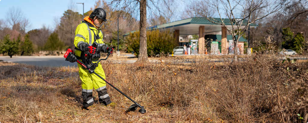 Opérateur  en tenue de protection jaune fluo débroussaillant des herbes sèches devant une station service avec une débroussailleuse Kress KC180.9 avec moteur arrière, la plus puissante de sa gamme
